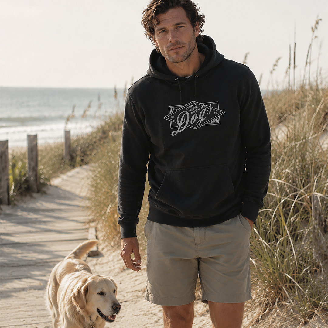 Man wearing a black hoodie with a logo and beige shorts walking a dog on a beach path.
