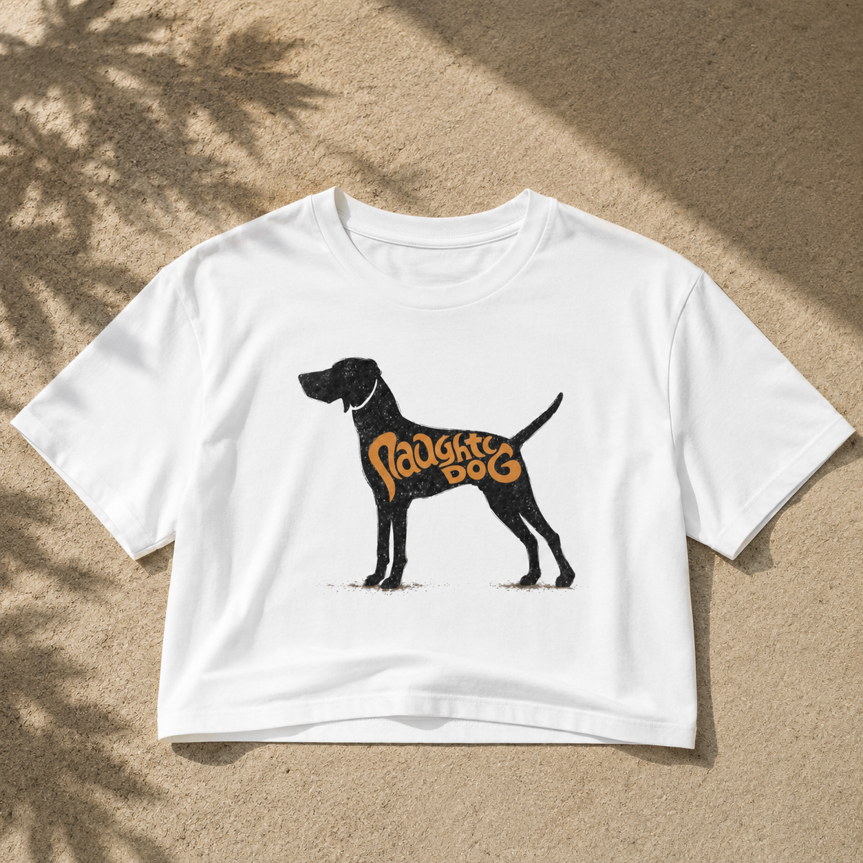 A white cropped short-sleeve T-shirt laid flat on a sandy surface with dappled shadows. The shirt features a black dog silhouette graphic with the words 'Puppy Dog' in orange across the torso.