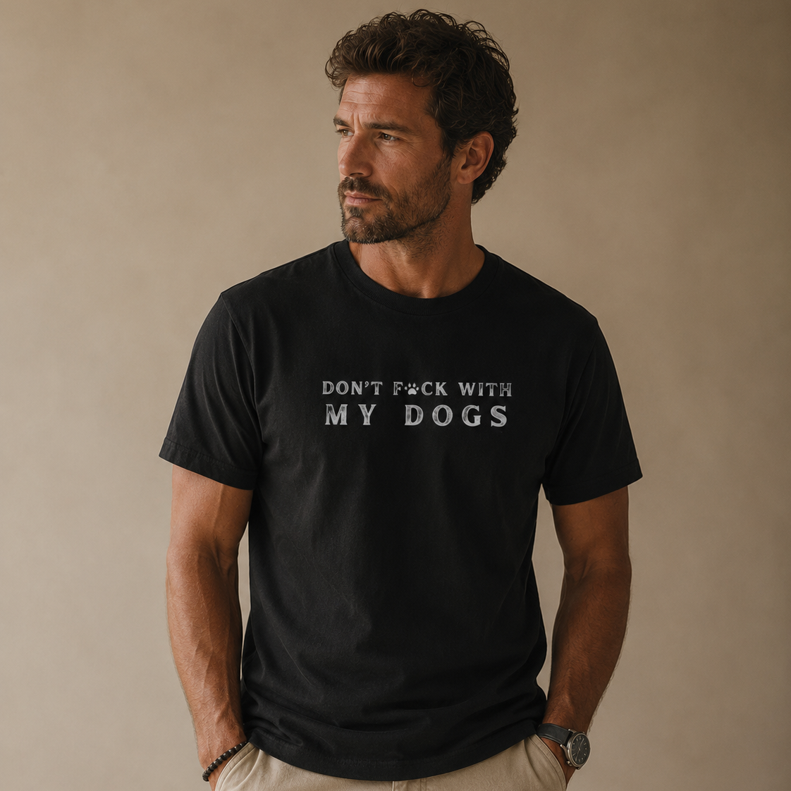 A man wearing a black crewneck t-shirt with the text 'DON'T F*CK WITH MY DOGS' printed in white across the chest, paired with khaki pants, standing against a neutral beige background.