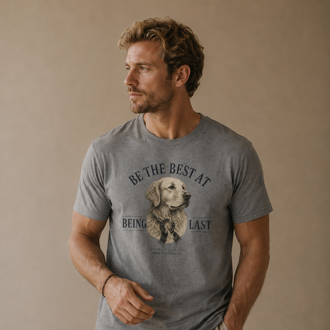 A man wearing a heather gray graphic t-shirt with a golden retriever illustration and the text 'BE THE BEST AT BEING LAST', paired with beige pants, set against a plain beige studio background.