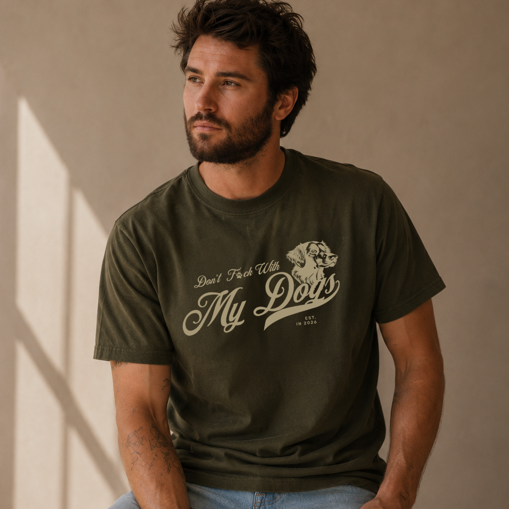 A man wearing an olive green graphic t-shirt with a cream-colored 'Don't Forget My Dog' print, paired with light-wash ripped jeans, sitting on a wooden stool against a textured beige wall.