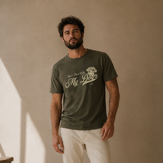 A man wearing an olive green graphic t-shirt with 'Don't Be My Dad' printed on the front, paired with off-white pants and white sneakers, standing in a warm-toned studio setting.