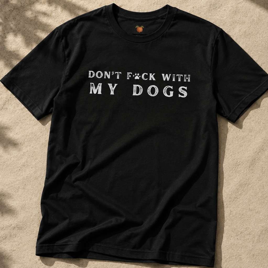 A black short-sleeve t-shirt with the text 'DON'T F*CK WITH MY DOGS' printed in white across the chest, laid flat on a sandy surface with dappled shadows.