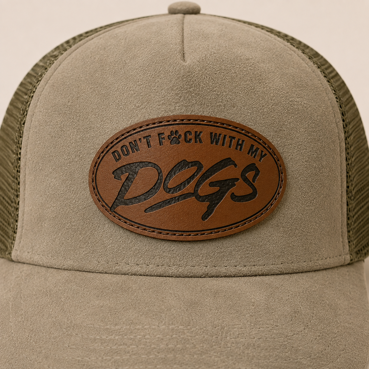 A close-up of a trucker hat with a beige suede front panel and olive green mesh back. A brown oval leather patch on the front is embossed with the text 'DON'T F*CK WITH MY DOGS'.