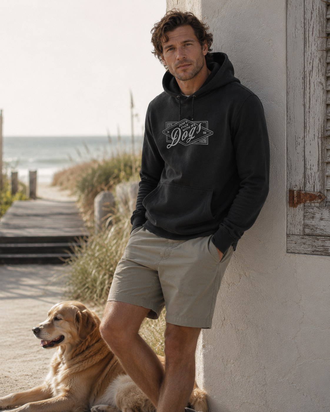 Man wearing a black hoodie with a logo and beige shorts standing next to a dog by a white wall.