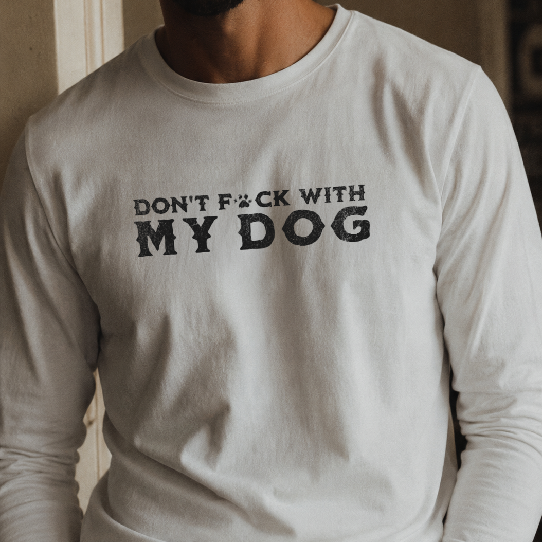 A man wearing a white long-sleeve t-shirt with the bold black text 'DON'T F*CK WITH MY DOG' printed across the chest, paired with dark blue jeans.