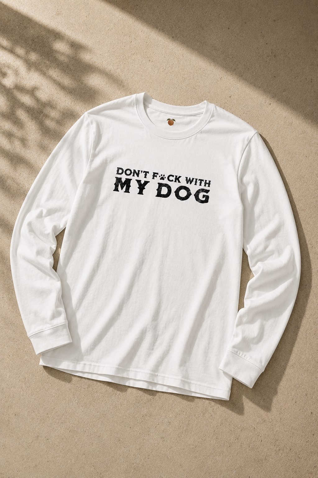 A white long-sleeve crewneck t-shirt with the text 'DON'T F*CK WITH MY DOG' printed in black across the chest, laid flat on a textured beige background with soft leaf shadows.