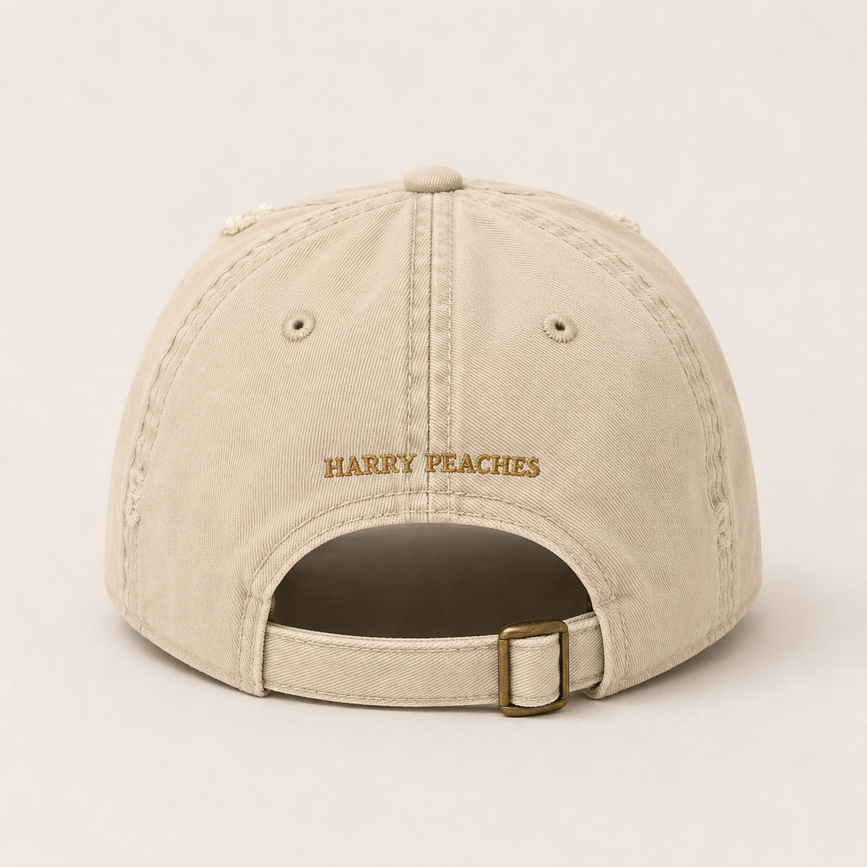 A beige baseball cap shown from the back, featuring 'HARRY PEACHES' embroidered in gold on the center back and an adjustable strap with a gold-tone buckle.