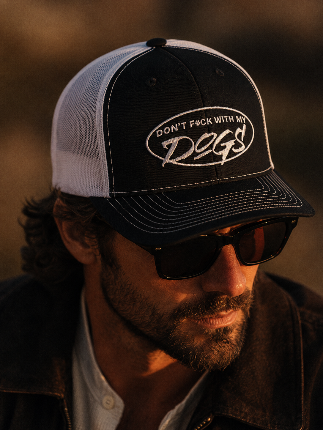 Man wearing a black and white cap with text, sunglasses, and a dark jacket against a blurred background.