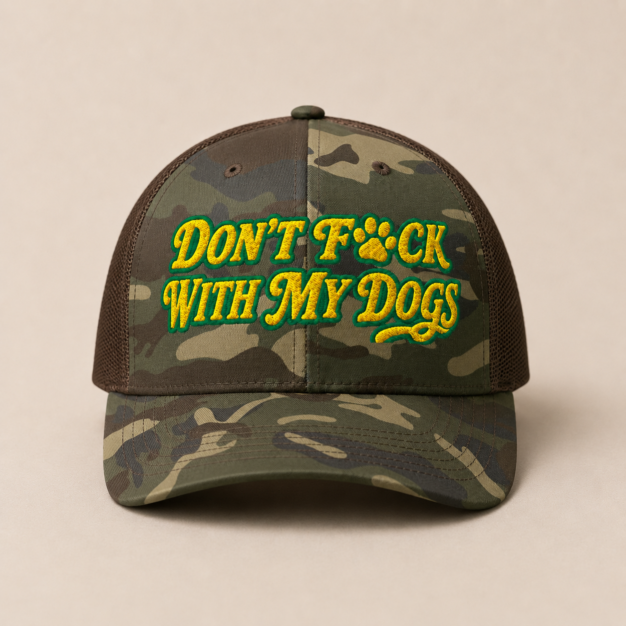 A camouflage trucker cap with brown mesh side panels and a curved brim, featuring the embroidered text 'DON'T F*CK WITH MY DOGS' in yellow with green outlines and a paw print graphic on the front. The cap is photographed against a solid, warm beige background.
