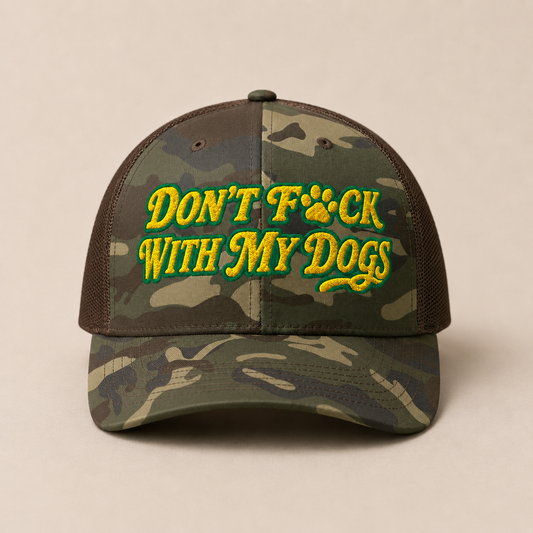 A camouflage trucker cap with brown mesh side panels and a curved brim, featuring the embroidered text 'DON'T F*CK WITH MY DOGS' in yellow with green outlines and a paw print graphic on the front. The cap is photographed against a solid, warm beige background.