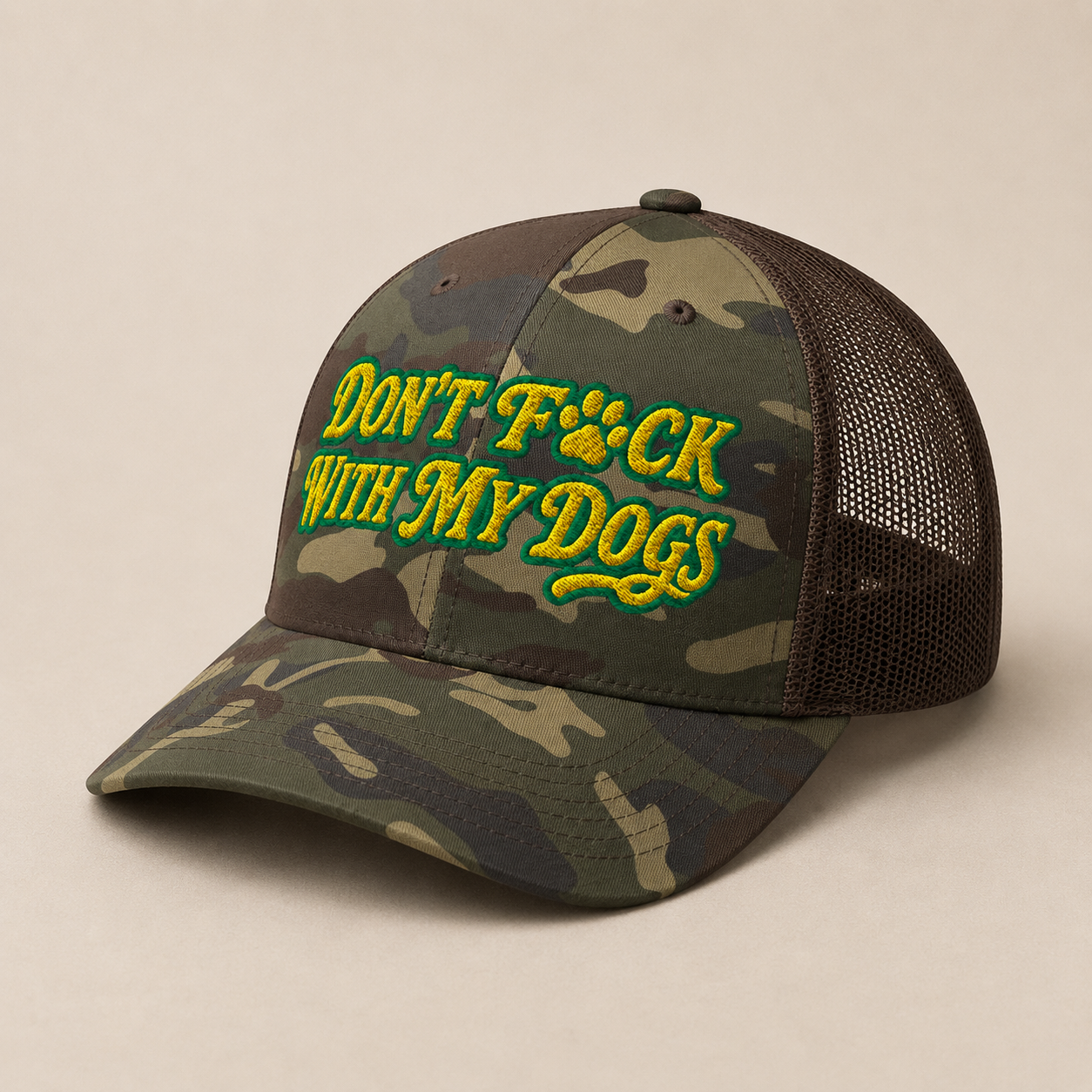 A camouflage trucker cap with dark brown mesh back panels and embroidered green text reading 'DON'T F*CK WITH MY DOGS' across the front. The cap is photographed on a plain beige background.