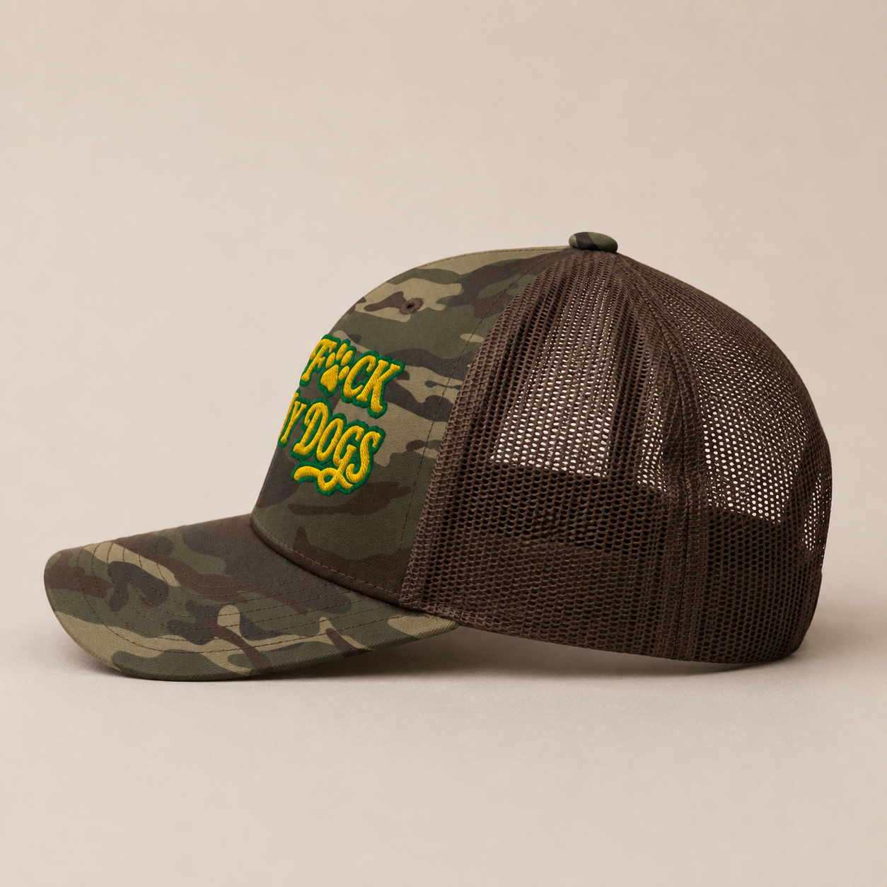 A camouflage trucker hat with a dark brown mesh back and green embroidered text reading 'TRUCKER DOGS' on the front panel, shown against a plain beige background.