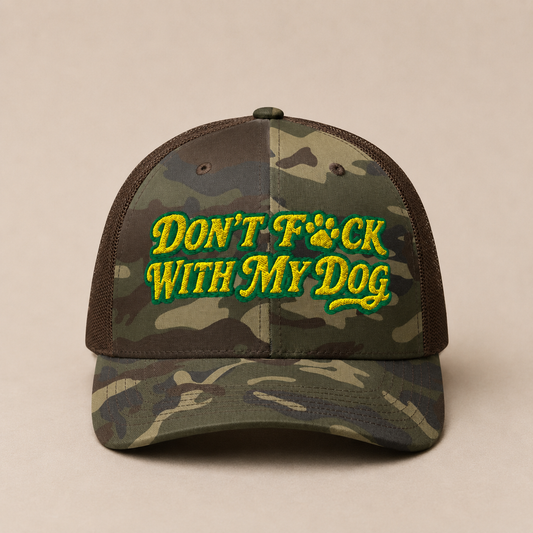 A camouflage trucker cap with brown mesh back panels and green and yellow embroidered text reading 'DON'T F*CK WITH MY DOG' on the front, shown against a plain beige background.