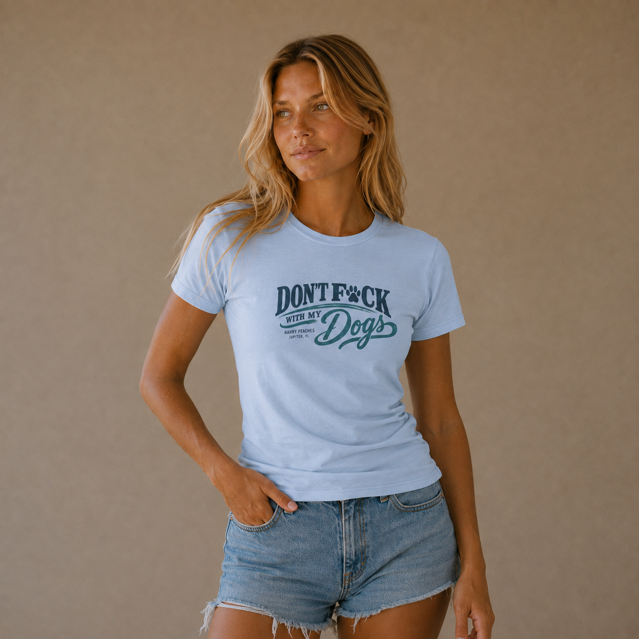 A woman modeling a light blue crewneck t-shirt with a dark teal graphic that reads 'DONT F*CK OUR DOGS' against a solid beige background.