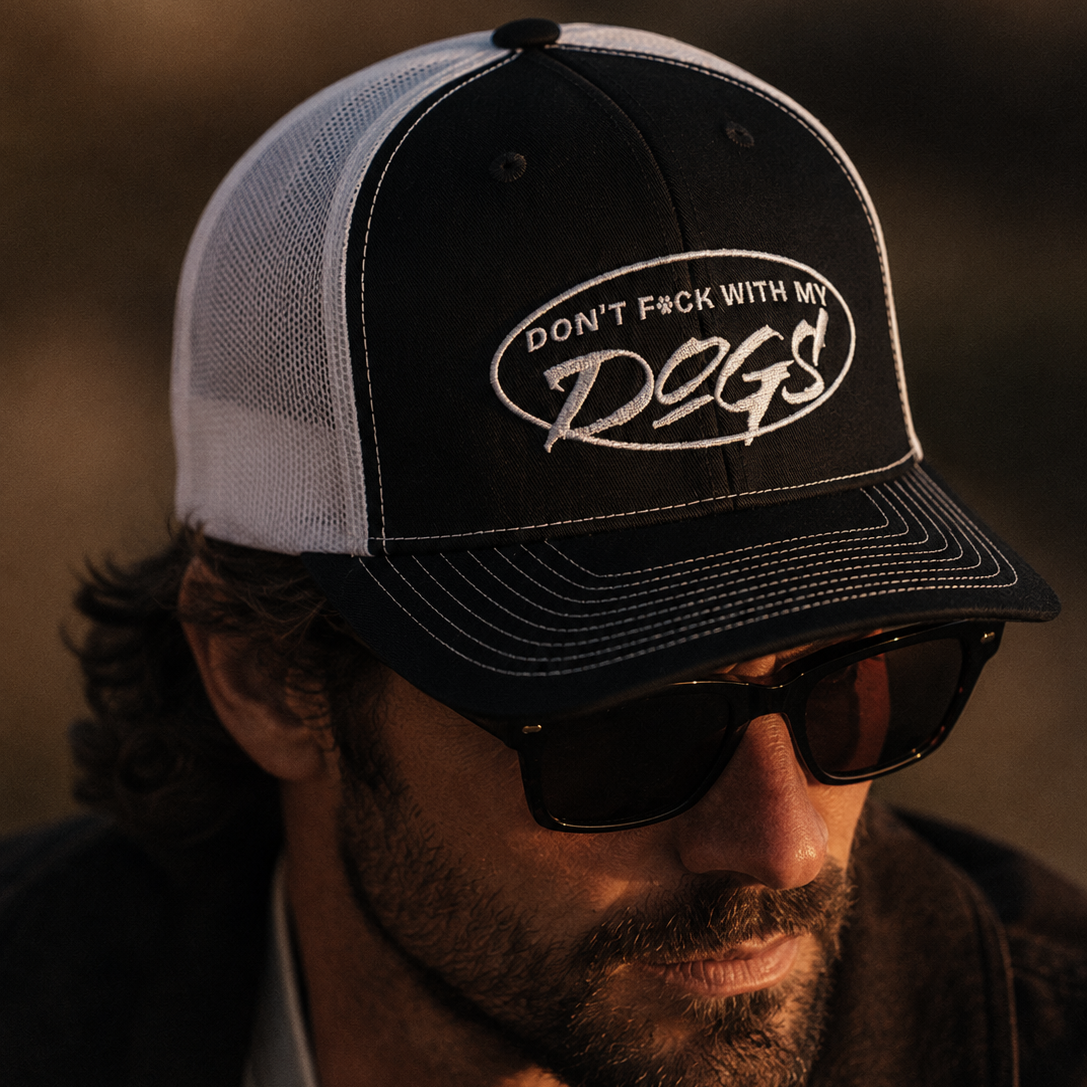 Man wearing a cap with 'Don't F**k with my dogs' text, sunglasses, and dark jacket.