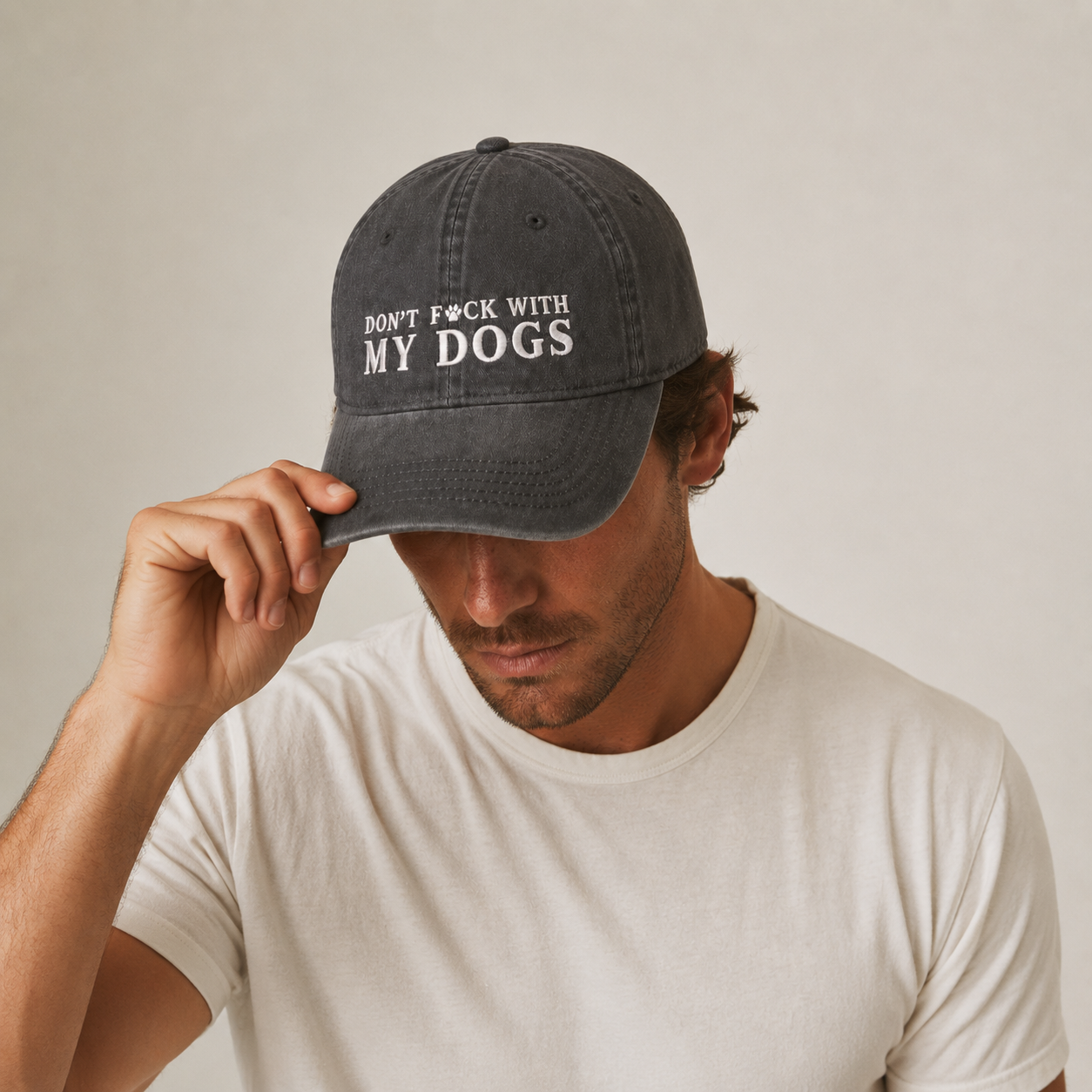 A man wearing a dark gray washed baseball cap with the white embroidered text 'DON'T F*CK WITH MY DOGS' and a white t-shirt, holding the brim of the cap with his hand against a plain light beige background.
