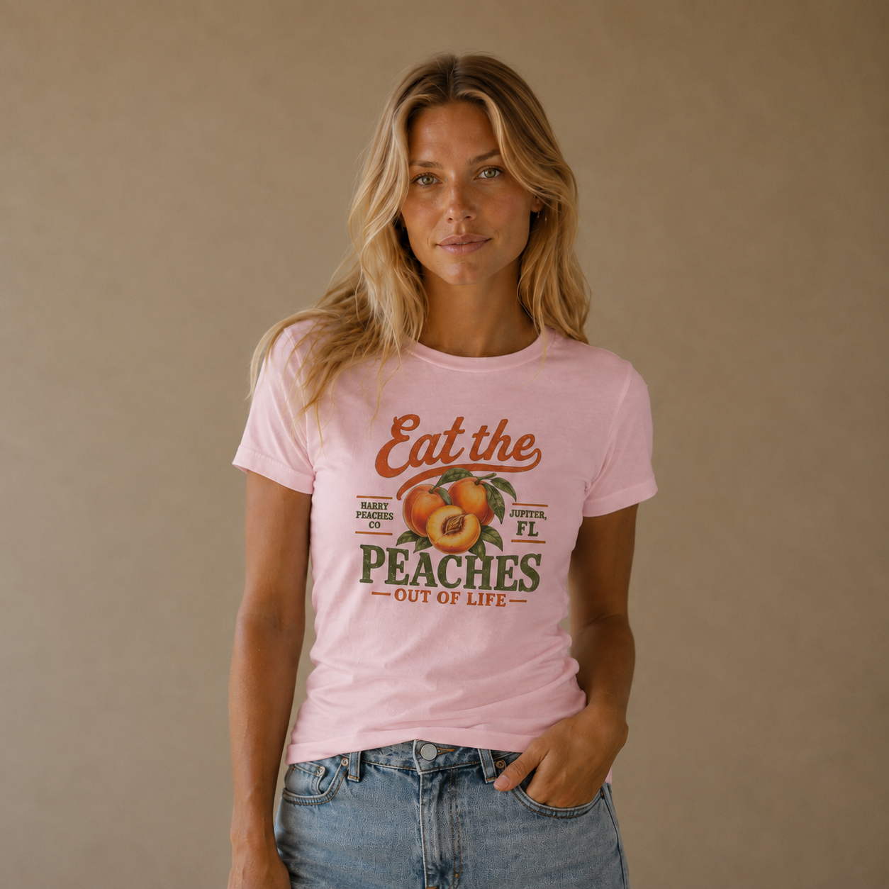 A woman wearing a light pink graphic t-shirt with a retro peach design and text reading 'Eat the PEACHES OUT OF LIFE', paired with blue jeans, standing against a solid beige background.