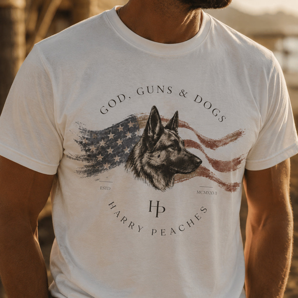 White t-shirt with a graphic of a dog, American flag, and text 'GOD, GUNS & DOGS' by Harry Peaches.