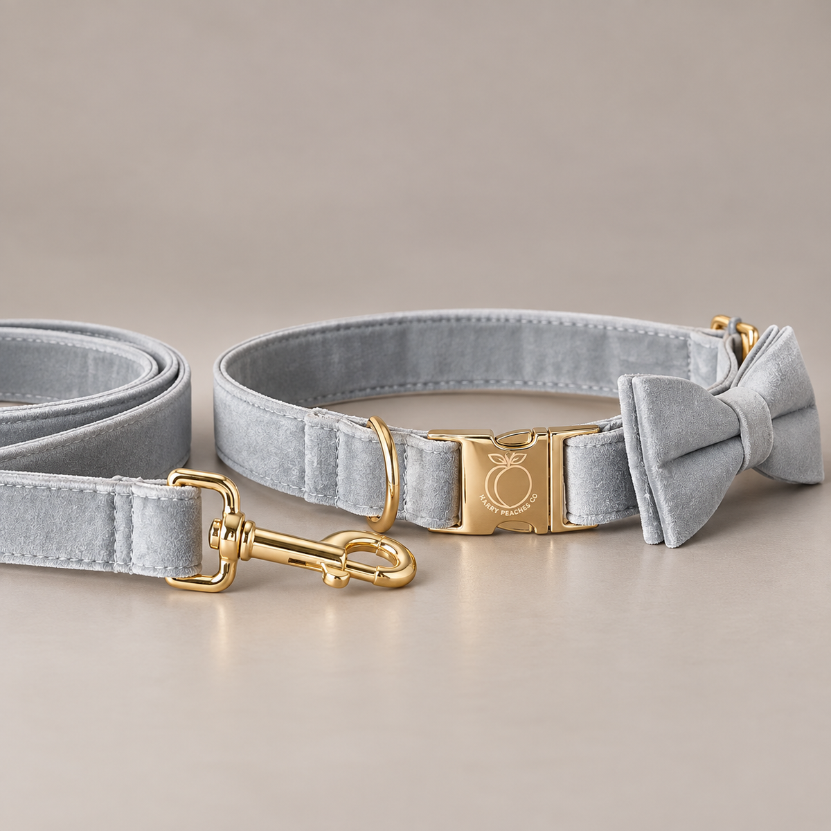 A light gray velvet pet collar with a matching bow tie and leash, featuring polished gold-tone hardware and a branded buckle, displayed on a neutral beige background.