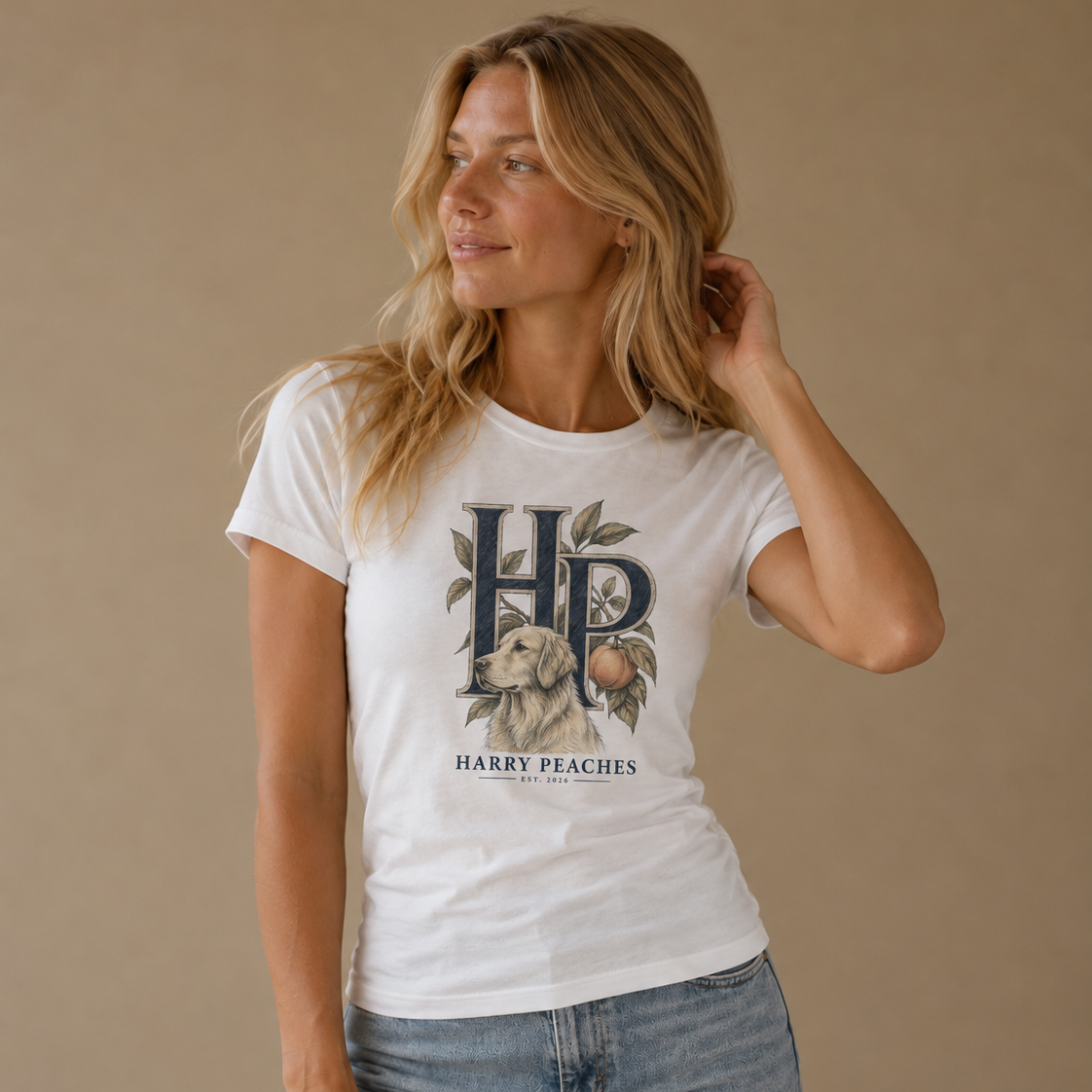 A woman modeling a white graphic t-shirt with a vintage-style 'HP' monogram, peach, and dog illustration, paired with light-wash blue jeans against a solid beige studio background.