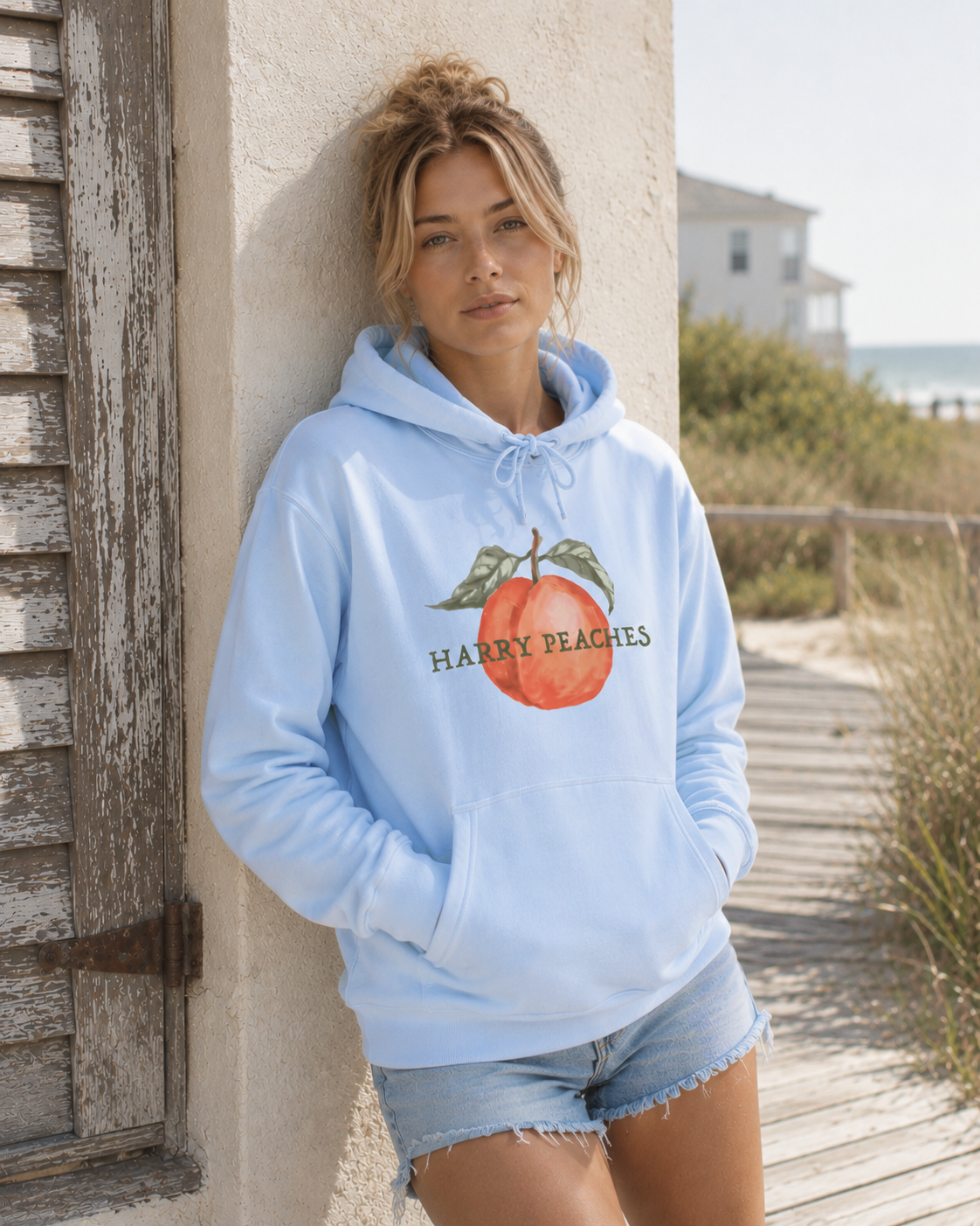 Woman wearing a light blue hoodie with 'Happy Peaches' graphic, standing by a wooden structure near a beach.