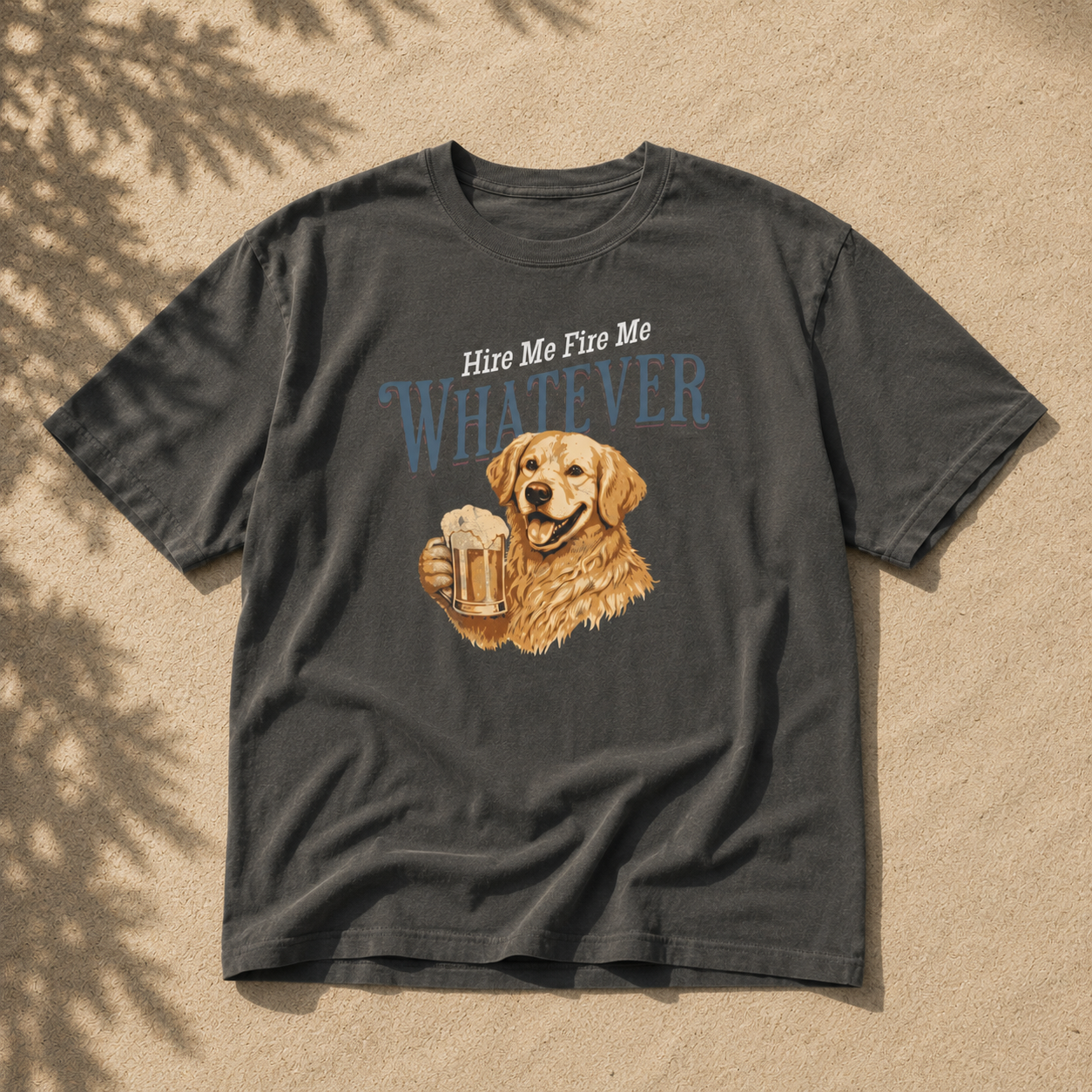 A dark charcoal gray graphic t-shirt laid flat on a sandy surface with leaf shadows. The shirt features a graphic of a golden retriever holding a beer mug, with the text 'Hire Me Fire Me Whatever' printed above.