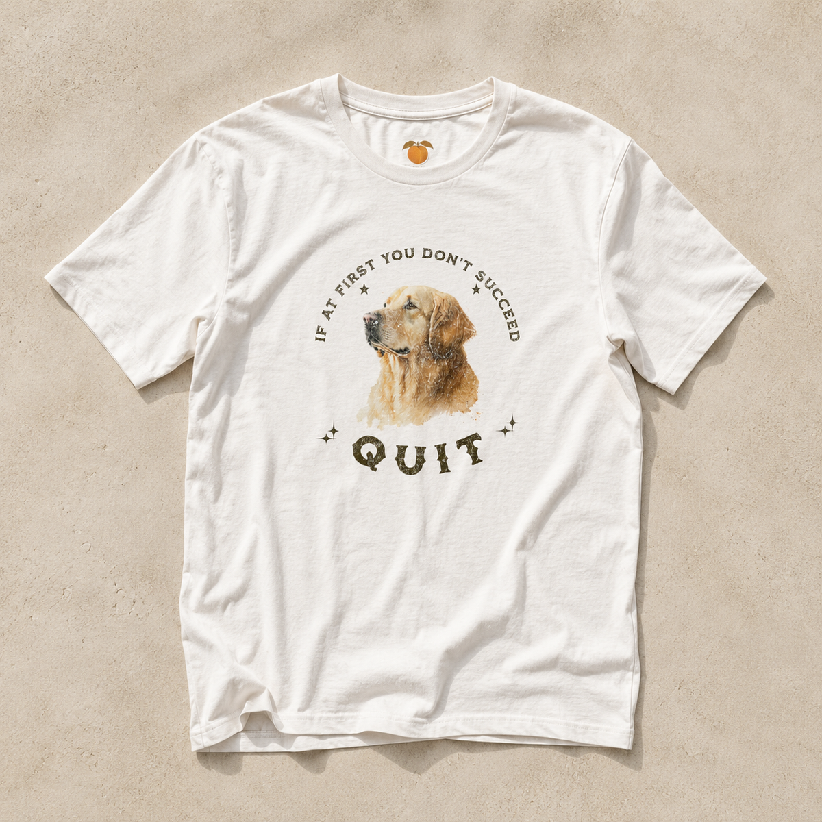 A white short-sleeve crewneck t-shirt laid flat on a textured beige background. The shirt features a graphic print of a golden retriever dog's face, surrounded by the text 'IF AT FIRST YOU DON'T SUCCEED QUIT' in a dark brown font.