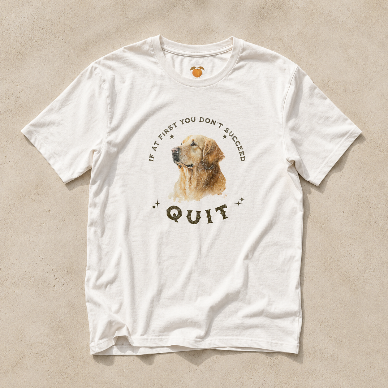 A white short-sleeve crewneck t-shirt laid flat on a textured beige background. The shirt features a graphic print of a golden retriever dog's face, surrounded by the text 'IF AT FIRST YOU DON'T SUCCEED QUIT' in a dark brown font.