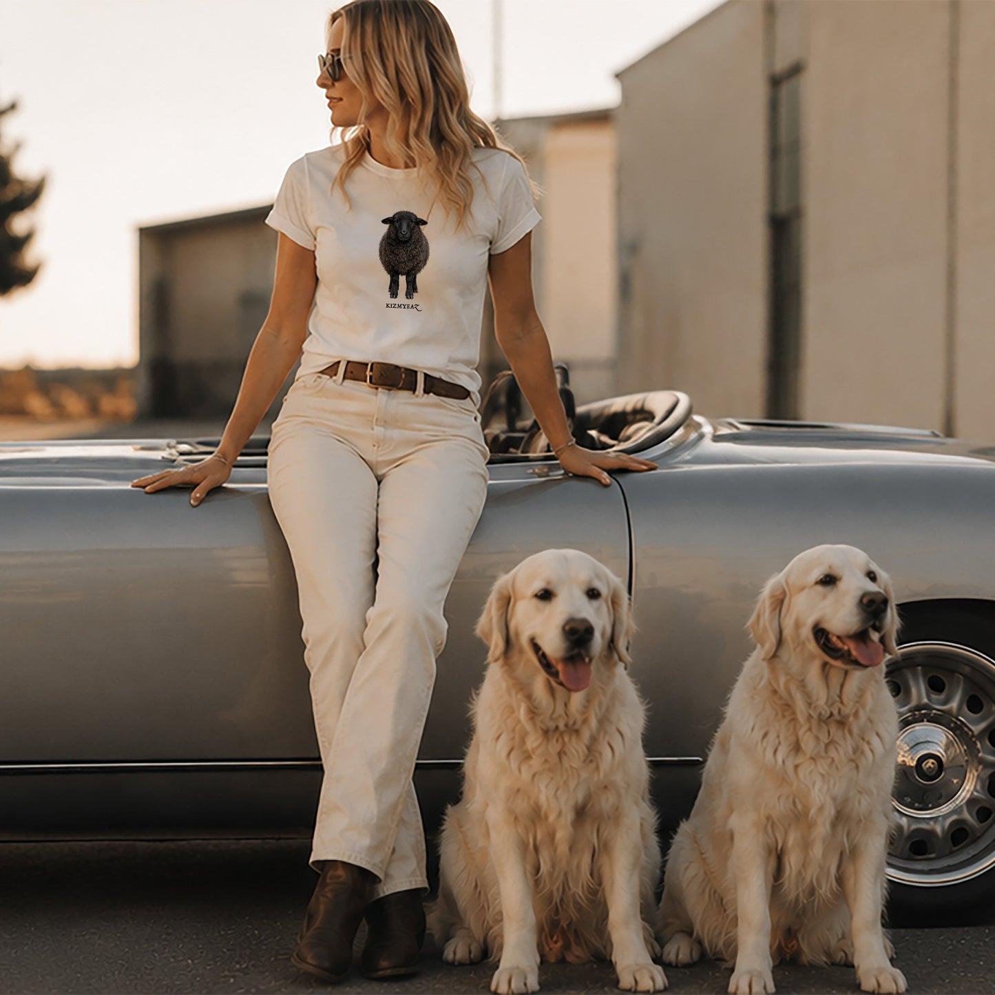 Woman sitting on a classic car with two dogs in front of a building.