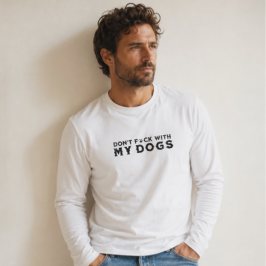 A man wearing a white long-sleeve t-shirt with the black text 'DON'T F*CK WITH MY DOGS' printed on the chest, paired with blue jeans, standing against a plain beige wall.