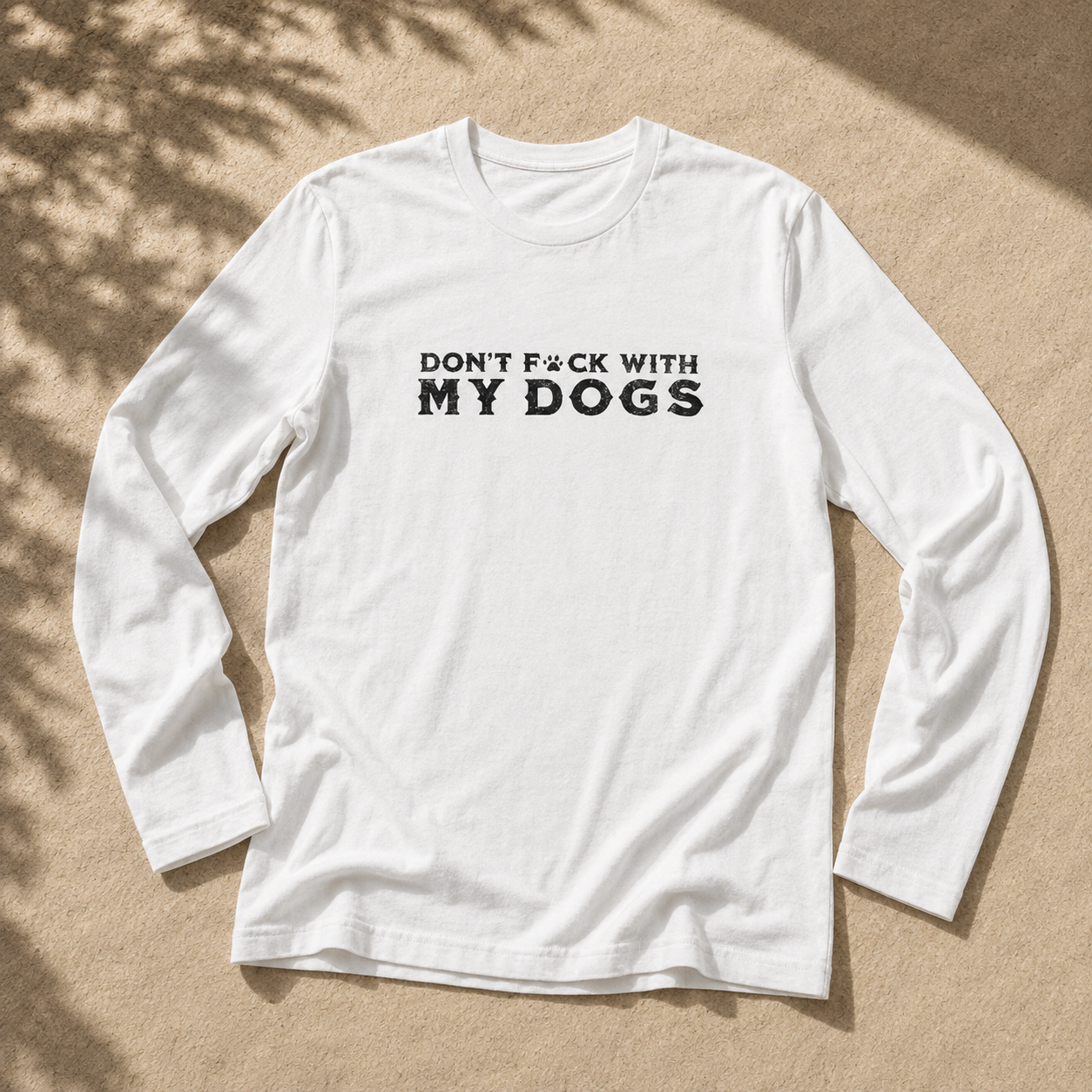A white long-sleeve t-shirt with the text 'DON'T F*CK WITH MY DOGS' printed in black across the chest, laid flat on a sandy surface with leaf shadows.