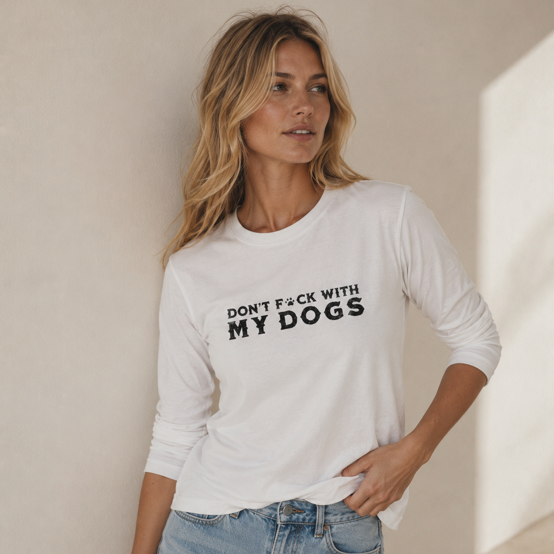 A woman wearing a white long-sleeve t-shirt with the black printed text 'DON'T F*CK WITH MY DOGS' and light-wash denim shorts, standing against a neutral beige wall.