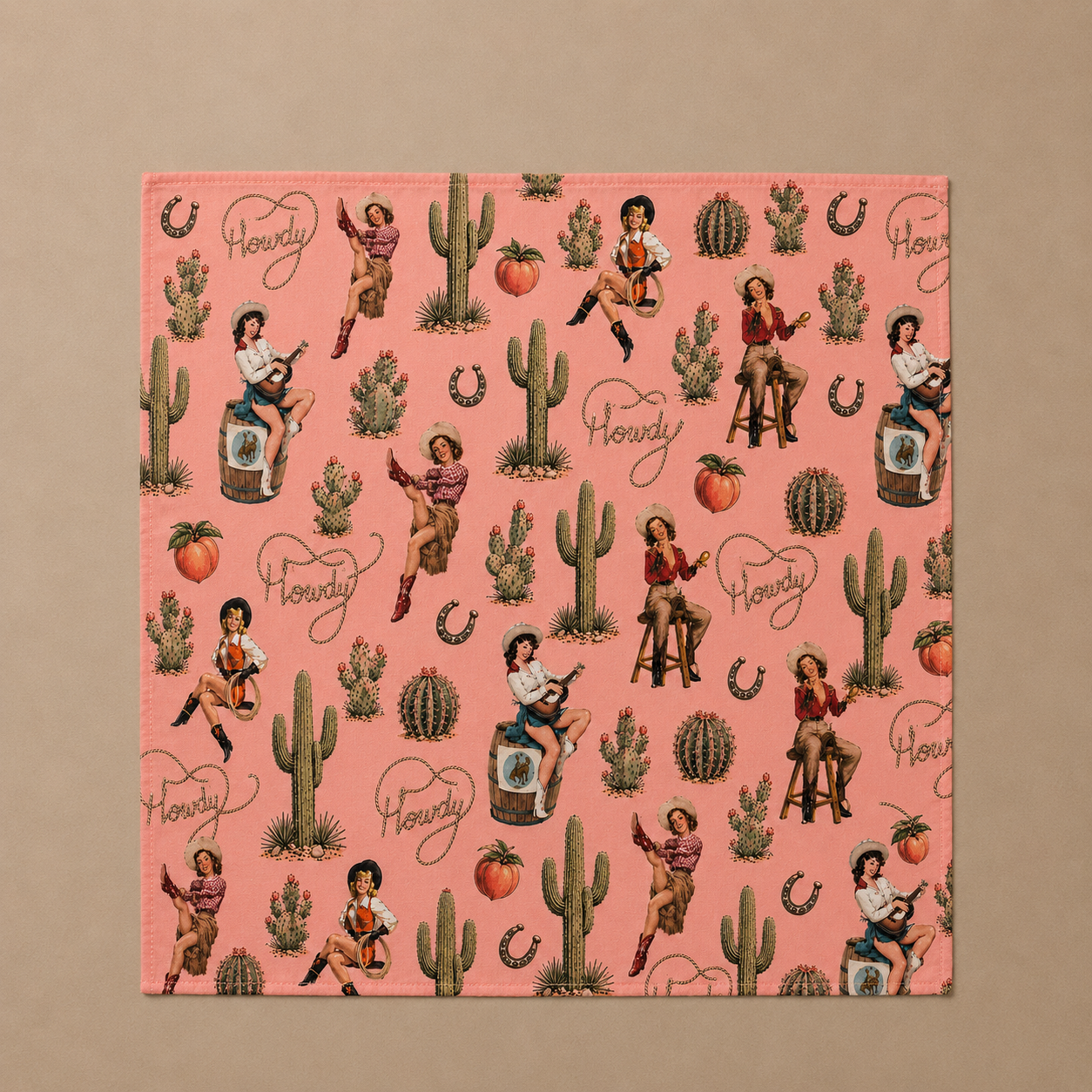 A square pink napkin featuring a vintage Western-themed print with cowboys, cacti, horseshoes, and the word 'Hollywood' repeated in script, displayed flat against a solid beige background.