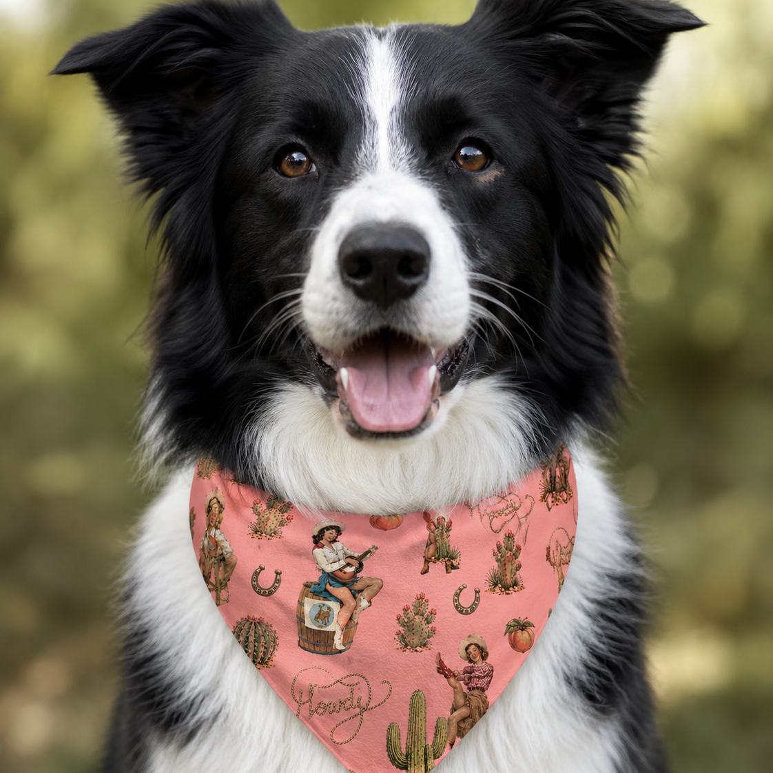 A black and white dog wearing a pink bandana with a western print featuring cacti, horseshoes, and cowboys, set against a soft-focus green outdoor background.