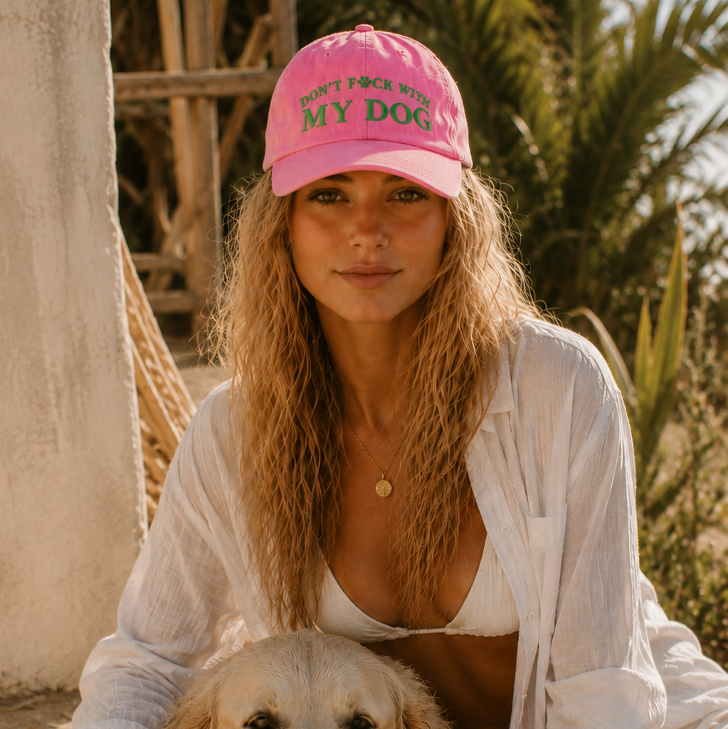 A woman wearing a pink baseball cap with green text that reads 'DON'T FORCE MY DOG INTO LEASH' sits outdoors with a golden retriever dog.