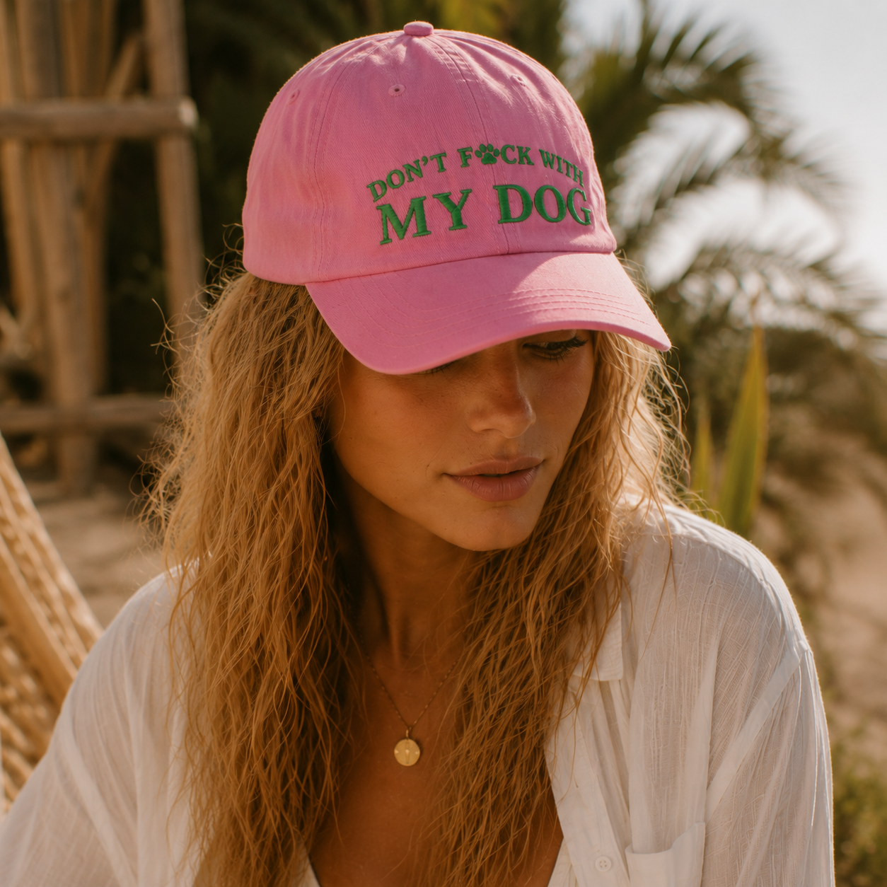 A woman wearing a pink baseball cap with green embroidered text reading 'DON'T F*CK WITH MY DOG' in an outdoor setting with warm sunlight and blurred palm trees in the background.