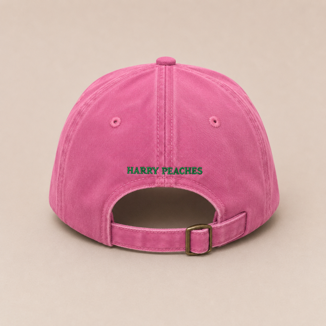 A pink cotton baseball cap shown from the back, featuring the text 'HARRY PEACHES' embroidered in green across the center. The cap has an adjustable strap with a metal buckle at the back and is photographed against a solid, warm beige background.