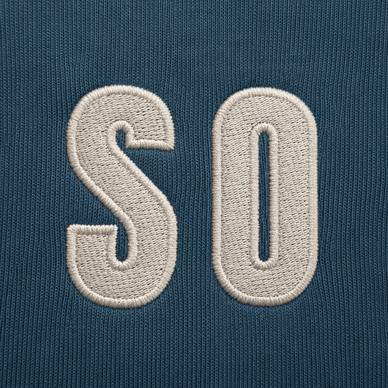 Close-up of a dark teal knit fabric with large cream embroidered letters 'SO' on the front.