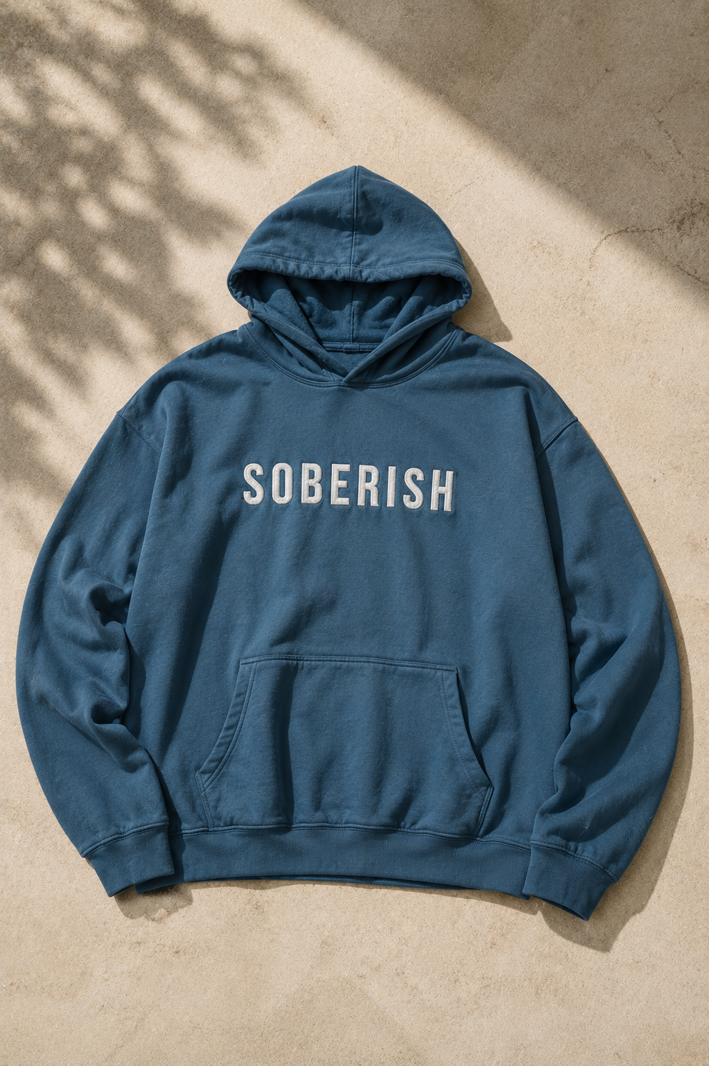 A dusty blue pullover hoodie with the word 'SOBERISH' printed in white across the chest, laid flat on a textured beige concrete surface with dappled sunlight and shadows.
