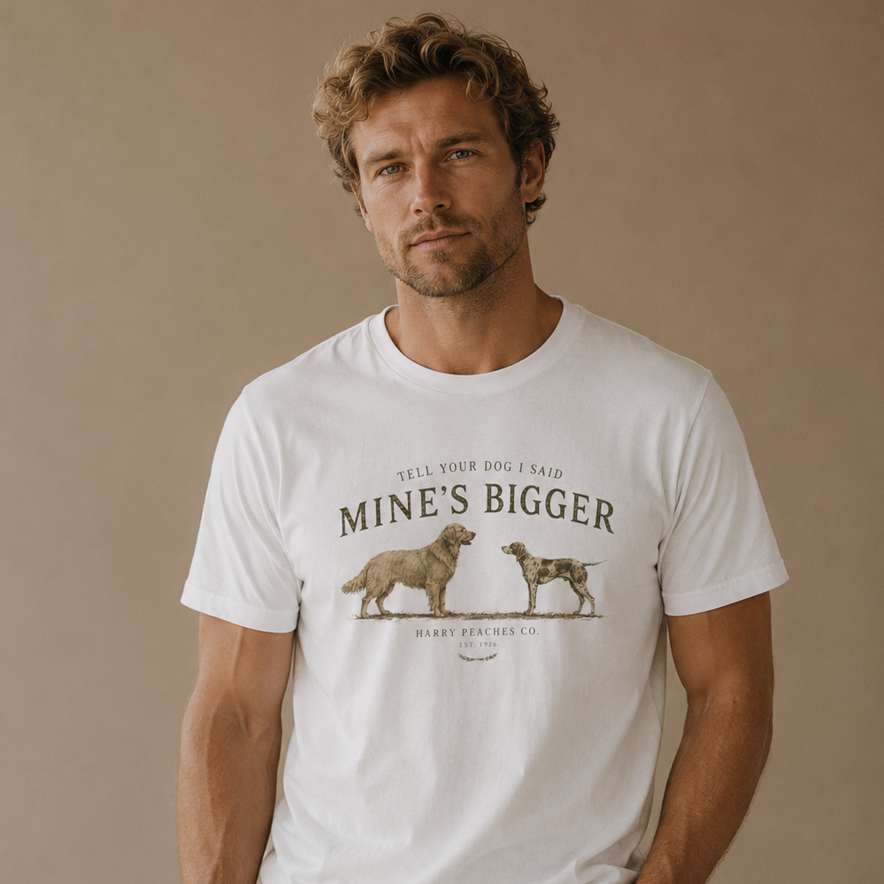 A man wearing a white graphic t-shirt with a brown print of two dogs and the text 'MINE'S BIGGER' and 'SELL YOUR DOG I SAID', paired with khaki pants, standing against a solid beige background.