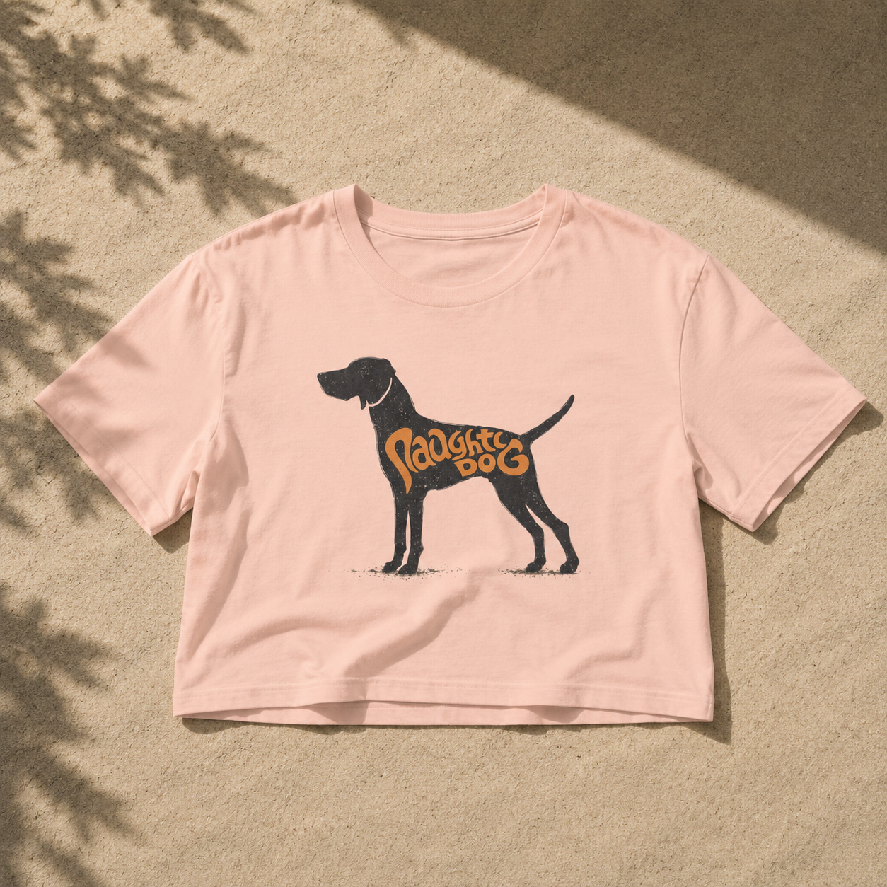 A light pink short-sleeve crop top t-shirt featuring a black dog silhouette graphic with the words 'Puppy Dog' in orange, laid flat on a sandy surface with dappled shadows.
