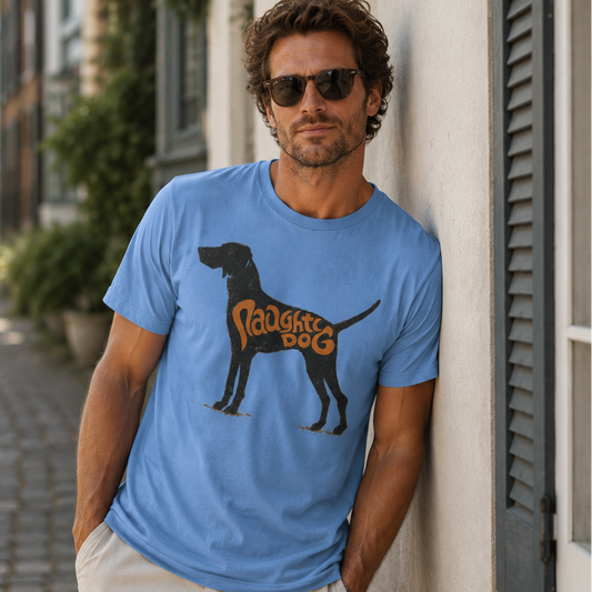A man wearing a light blue graphic t-shirt with a black dog silhouette and the words 'Proudly Dog' in orange, paired with beige shorts, standing on a cobblestone street.