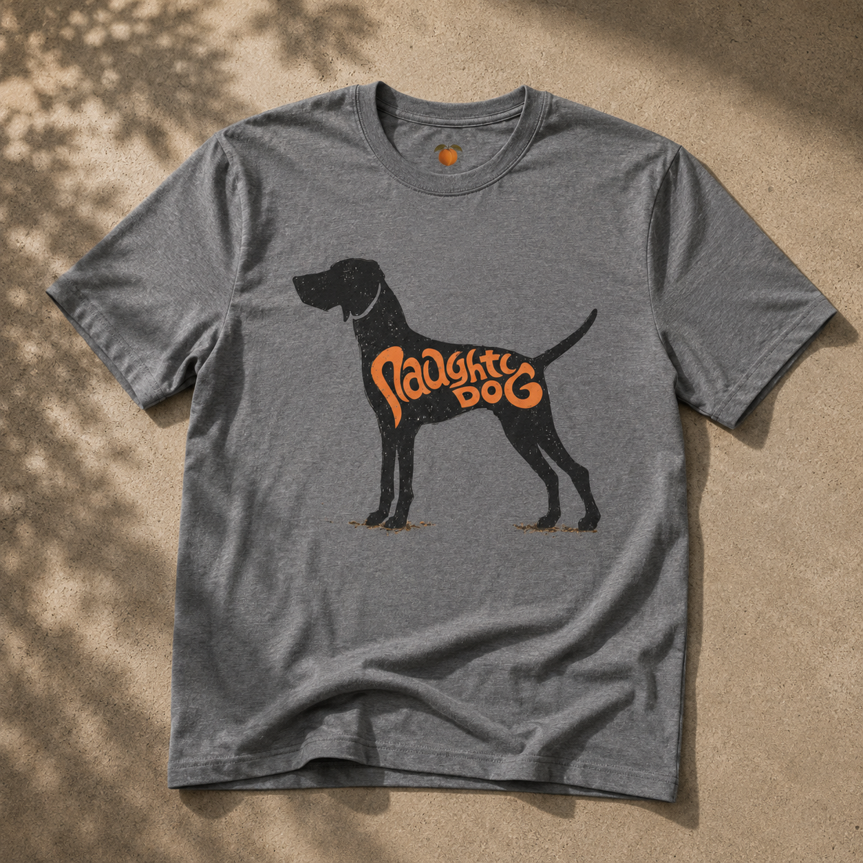 A heather gray short-sleeve t-shirt laid flat on a sandy surface with dappled shadows. The shirt features a black silhouette of a dog with the words 'Naughty Dog' printed in orange across its side.