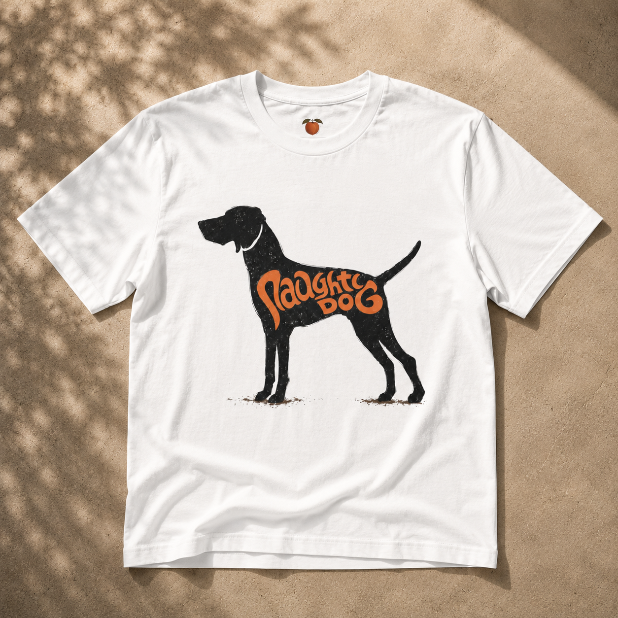A white short-sleeve t-shirt laid flat on a sandy surface with dappled shadows. The shirt features a black silhouette of a dog with the words 'Naughty Dog' printed in orange across its side.