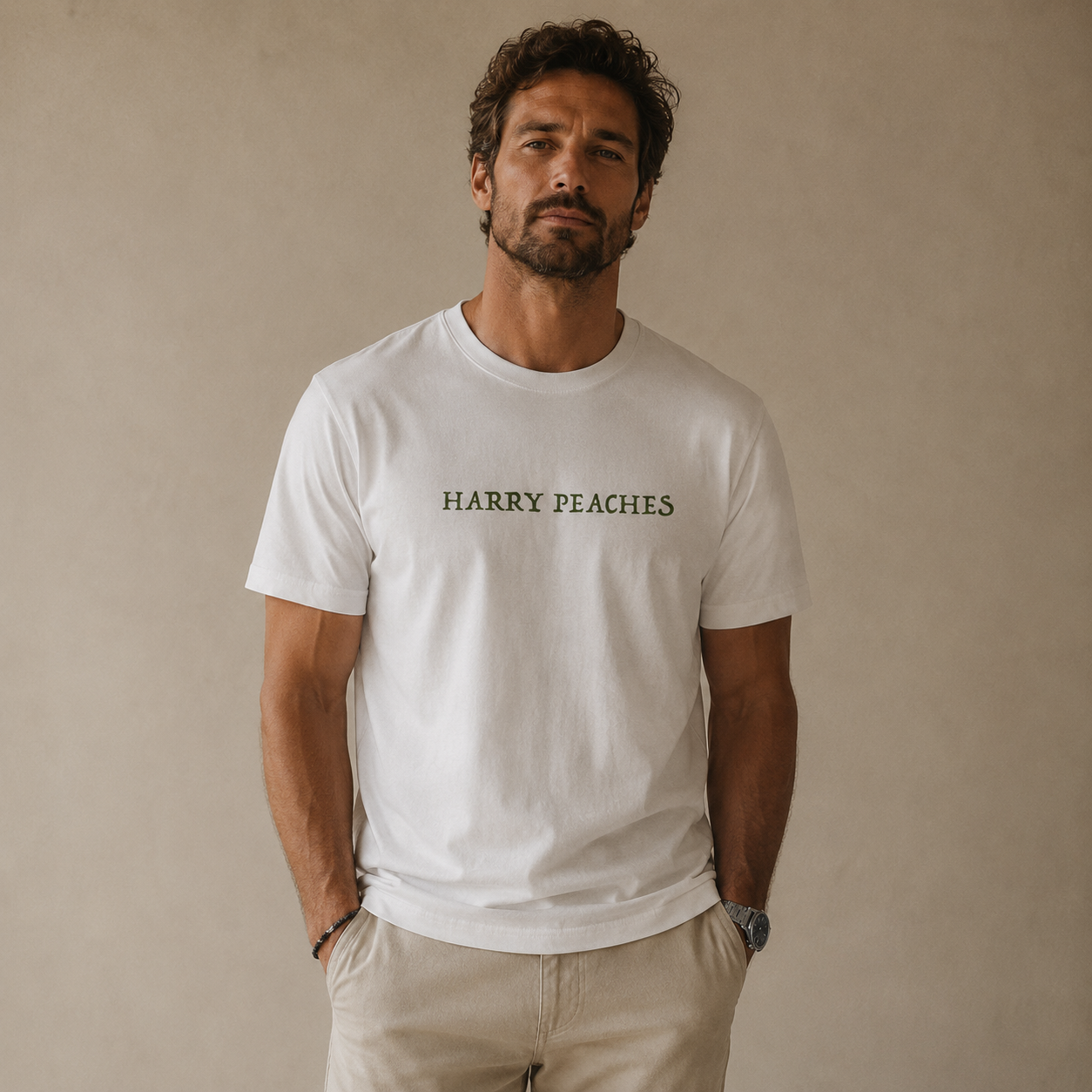 A man wearing a white crewneck t-shirt with 'HARRY PEACHES' printed in dark green across the chest, paired with beige trousers, standing against a neutral beige wall.