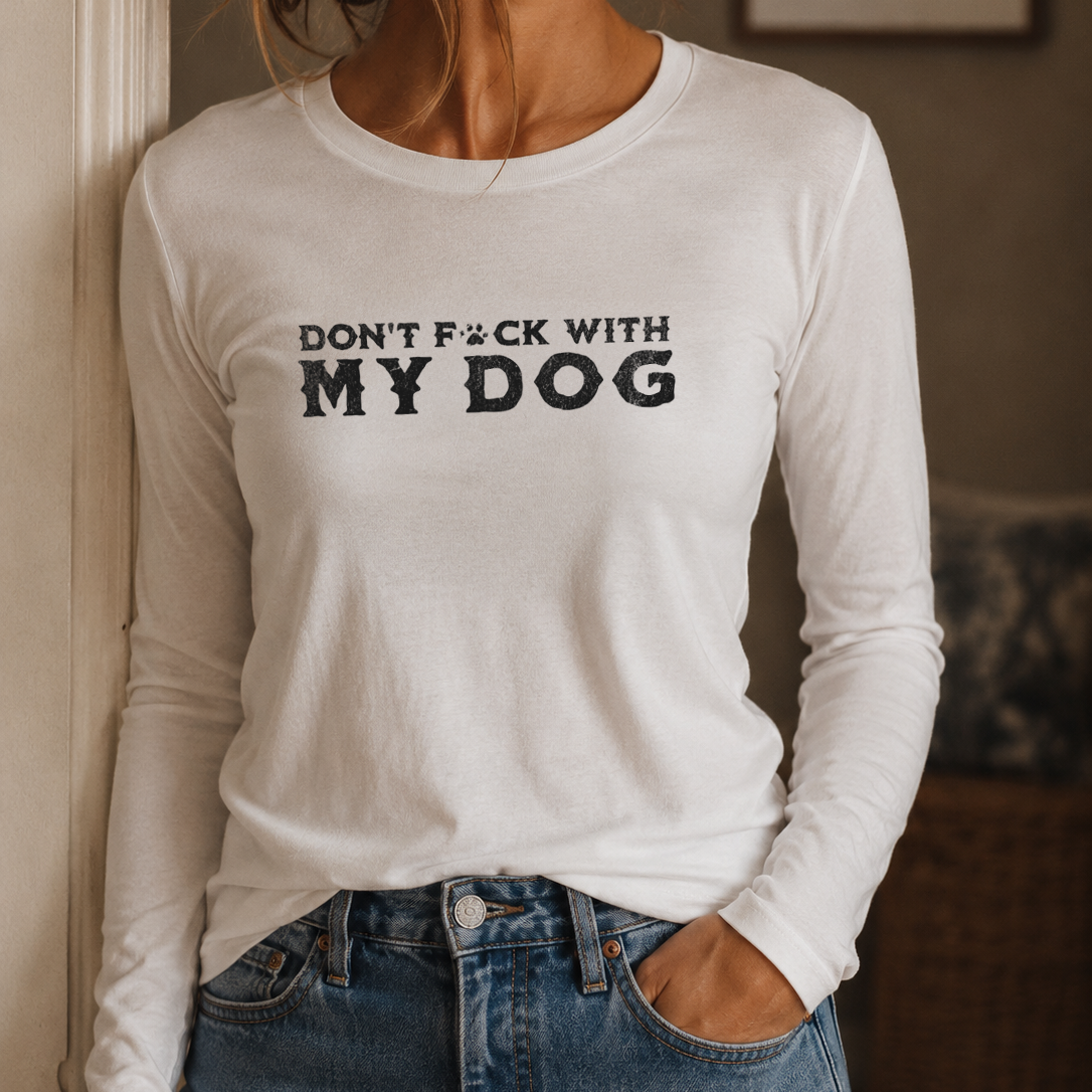 A woman wearing a white long-sleeve t-shirt with the text 'DON'T F*CK WITH MY DOG' printed in black across the chest, paired with blue jeans in an indoor setting.