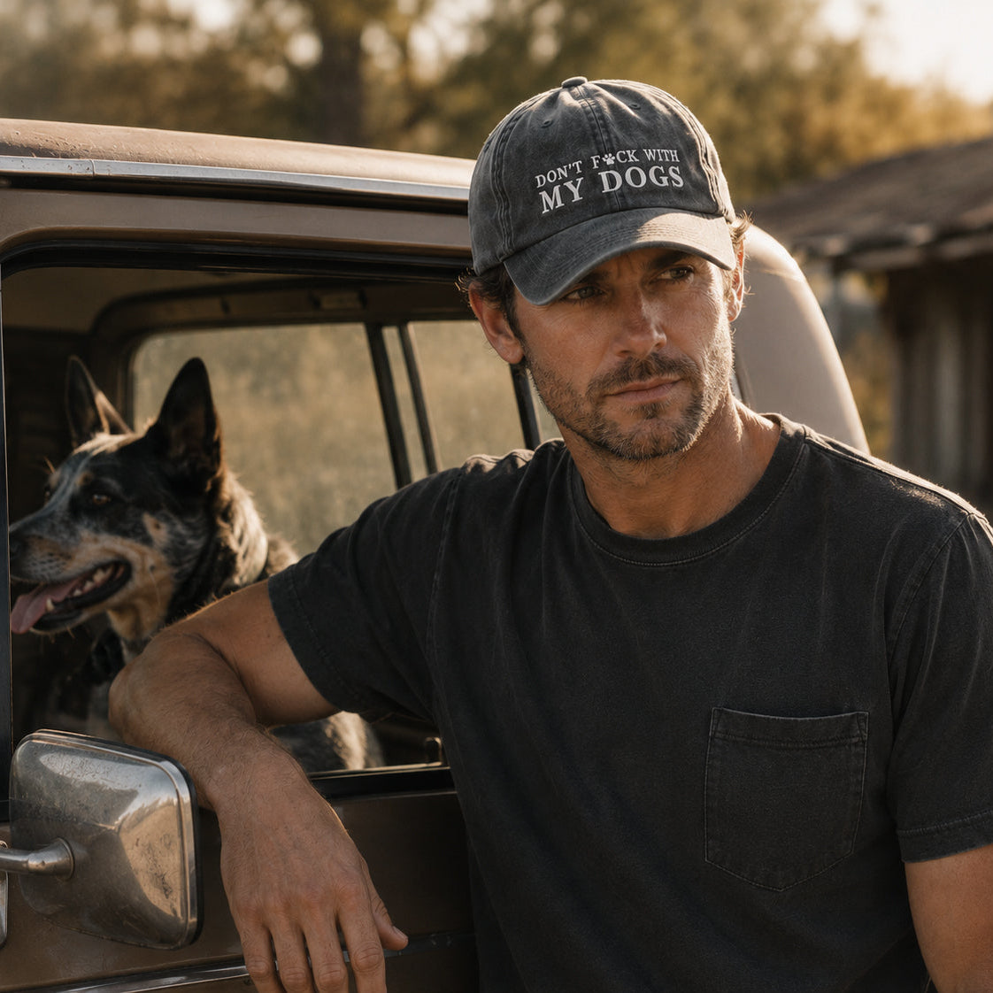 A man leaning out of a vintage pickup truck with a dog inside, wearing a dark gray t-shirt and a dark gray baseball cap that reads 'DON'T TACK WITH MY DOGS'.