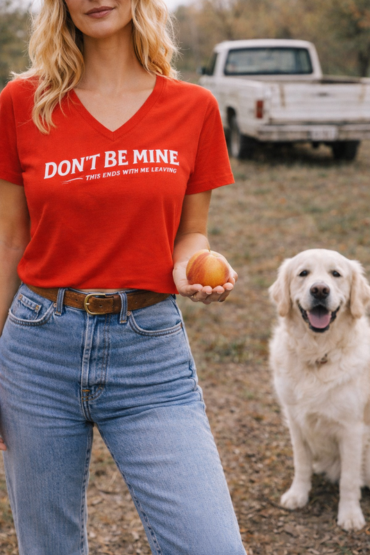 Person wearing a red t-shirt with text, holding an apple, standing next to a dog outdoors.