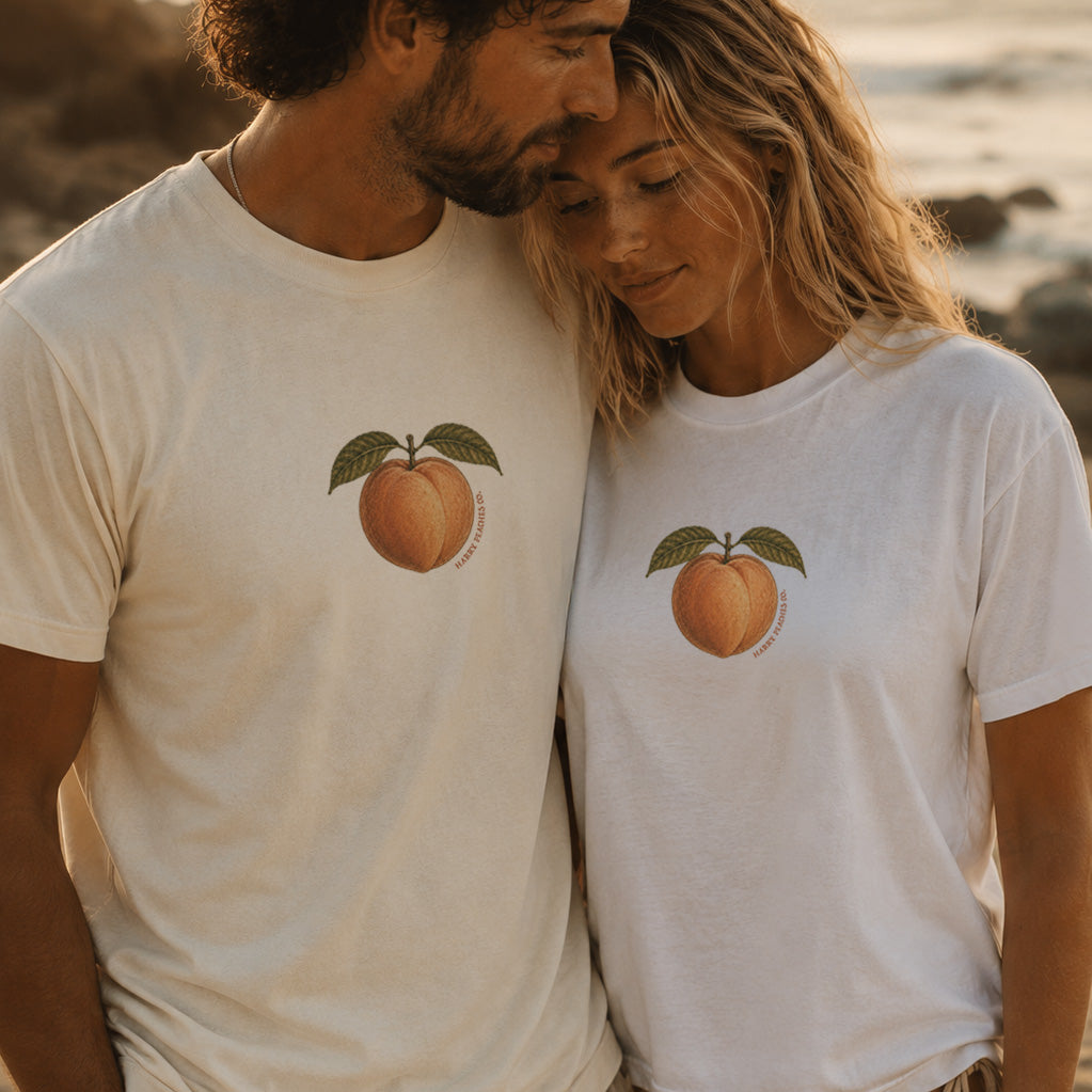 Two people wearing white t-shirts with peach graphics on a beach.